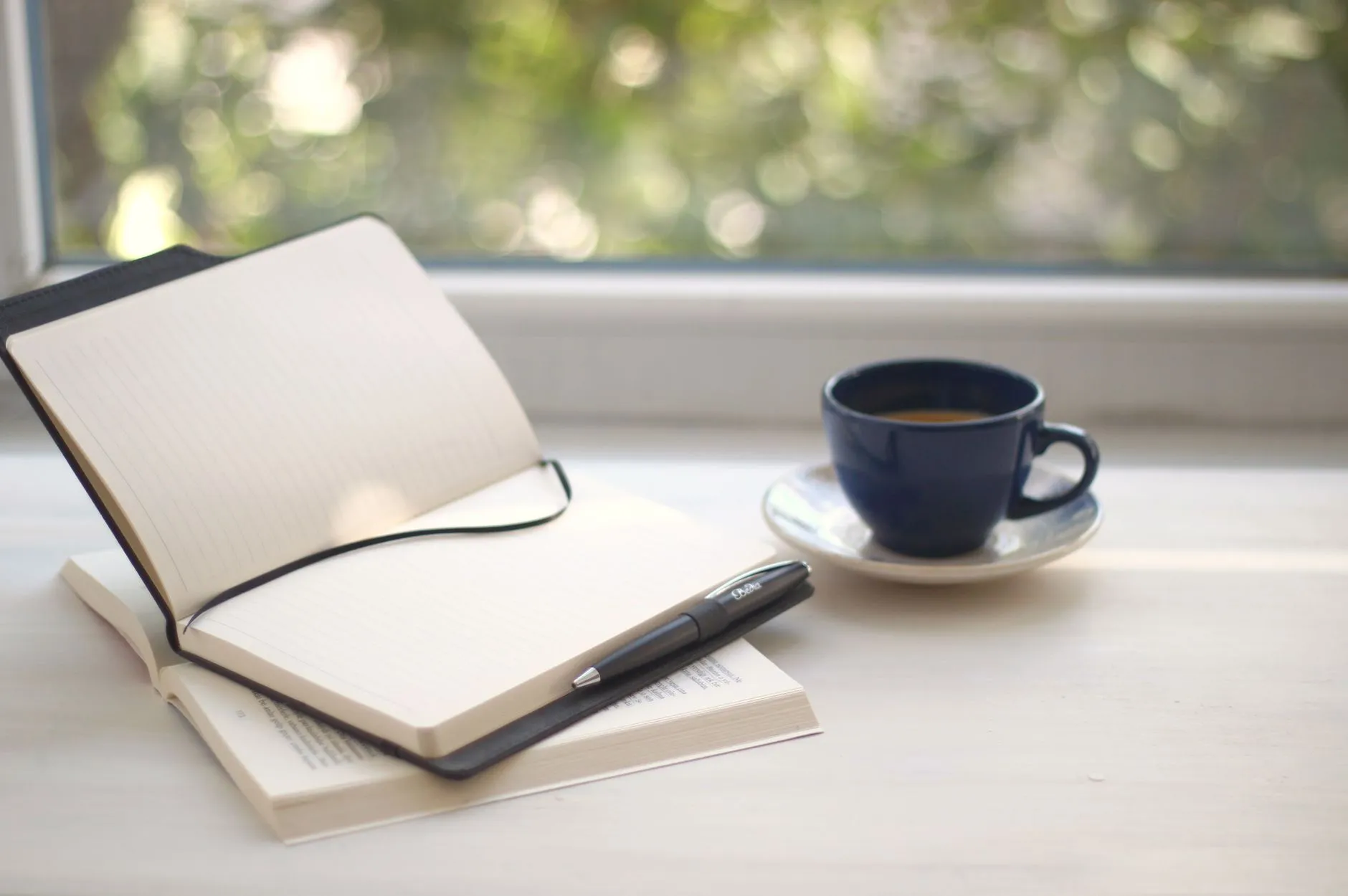 An open notebook, pen and cup of coffee on a sunlit windowsill